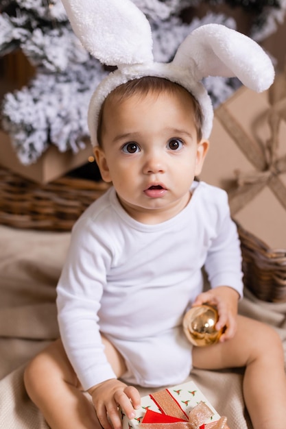 Schattige baby in een witte bodysuit en konijnenoren zit thuis naast de kerstboom en kijkt naar cadeaus symbool van het jaar het jaar van het konijn 2023