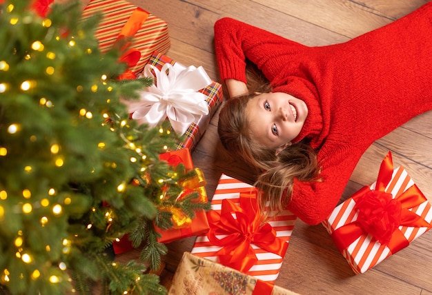 Schattige baby meisje ligt op de vloer bij de kerstboom met geschenken en kijkt naar de camera met een glimlach en vreugde thuis. Bovenaanzicht