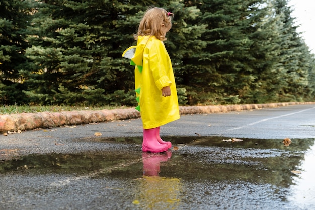 Schattige baby meisje peuter dragen gele stijlvolle regenjas roze rubberen laarzen staan in een plas.