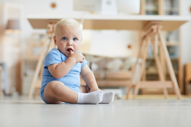 Schattige baby met blond haar en heldere blauwe ogen, zittend op de vloer met vinger in de mond