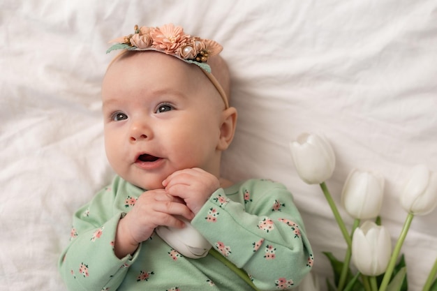 Schattige baby met een bloemenhoofdband en een groene katoenen bodysuit thuis op een wit bed met tulpen. Spr