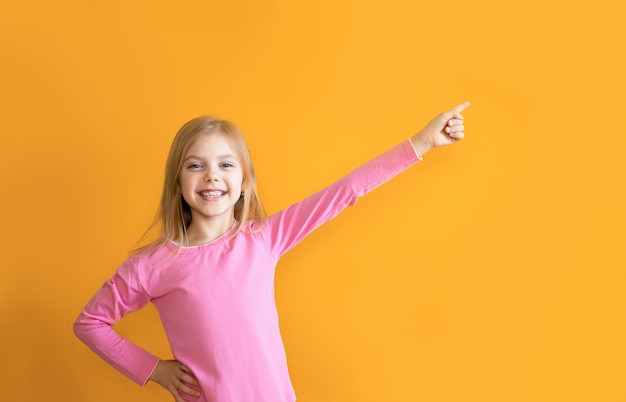 Schattige baby op een oranje muur, 6-8 jaar oud, een meisje in roze kleren glimlacht, wijst naar een specifieke plaats