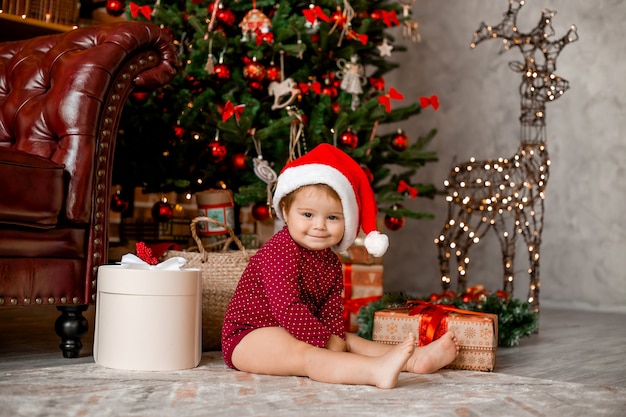 Schattige baby Santa zit thuis in de buurt van de kerstboom met geschenken