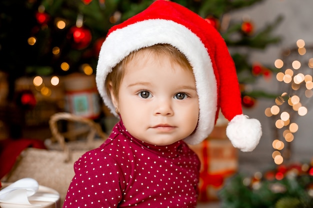 Schattige baby Santa zit thuis in de buurt van de kerstboom met geschenken