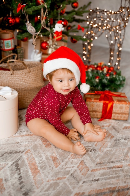 Schattige baby Santa zit thuis in de buurt van de kerstboom met geschenken
