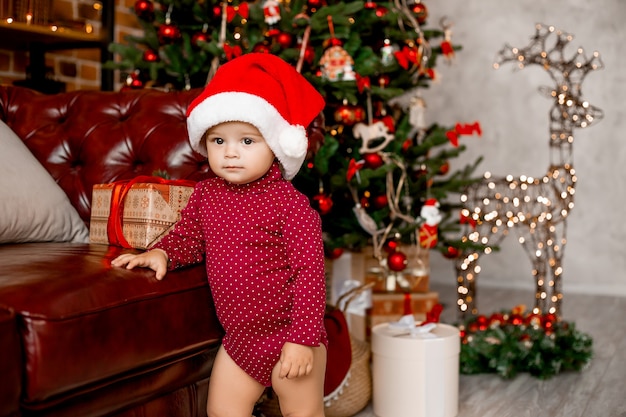 Schattige baby Santa zit thuis in de buurt van de kerstboom met geschenken