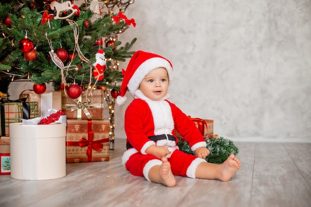 Schattige baby Santa zit thuis in de buurt van de kerstboom met geschenken