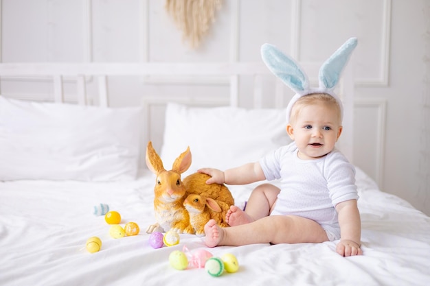 Schattige babyjongen met konijnenoren en kleurrijke eieren op een wit bed thuis kleine blonde baby spelen met een konijn gelukkig paasconcept