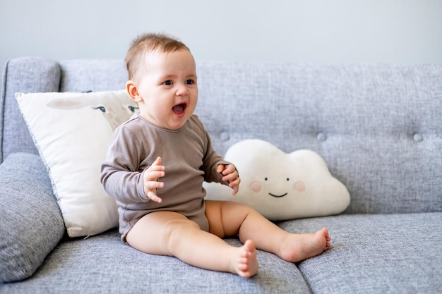 Schattige babyjongen van zes maanden speelt thuis op een grijze bank en lacht of lacht een gelukkige pasgeborene