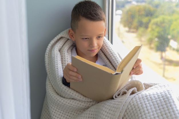 Schattige babyjongen zit bij het raam en leest een boek in de kamer thuis prachtige herfstnatuur