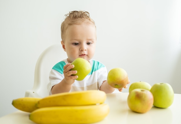 Schattige babyjongen zittend aan de tafel in kinderstoel appel eten op witte keuken.
