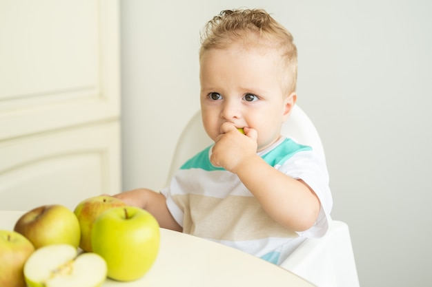 Schattige babyjongen zittend aan de tafel in kinderstoel appel eten op witte keuken.