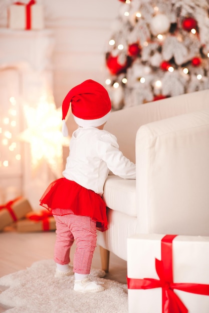 Schattige babymeisje 1 jaar oud dragen kerstmuts poseren over Kerstmis achtergrond. Achteraanzicht. Vakantie seizoen.