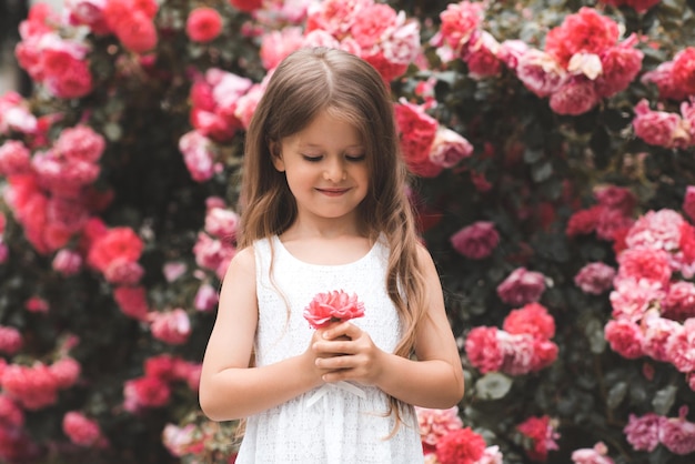 Schattige babymeisje 45 jaar oud met lang blond haar poseren over roze roze bloemen op de achtergrond close-up Lente glimlachend kind over bloeiende struiken in de tuin zomerseizoen geluk