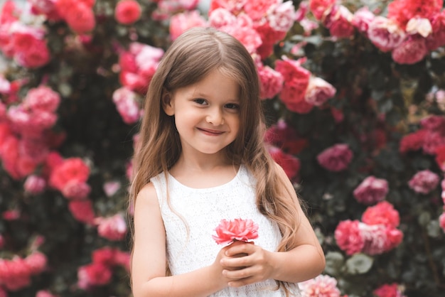 Schattige babymeisje 45 jaar oud met lang blond haar poseren over roze roze bloemen op de achtergrond close-up Lente glimlachend kind over bloeiende struiken in de tuin zomerseizoen geluk