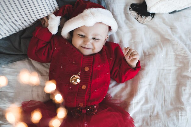 Schattige babymeisje draagt rode kerstmuts en gebreide jurk liggend in bed