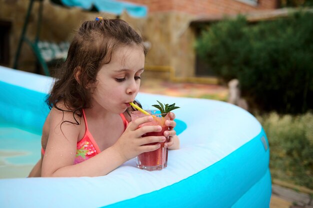 Schattige babymeisje drinkt vers geperst aardbeiensap terwijl ze ontspant in het zwembad op een warme zomerdag