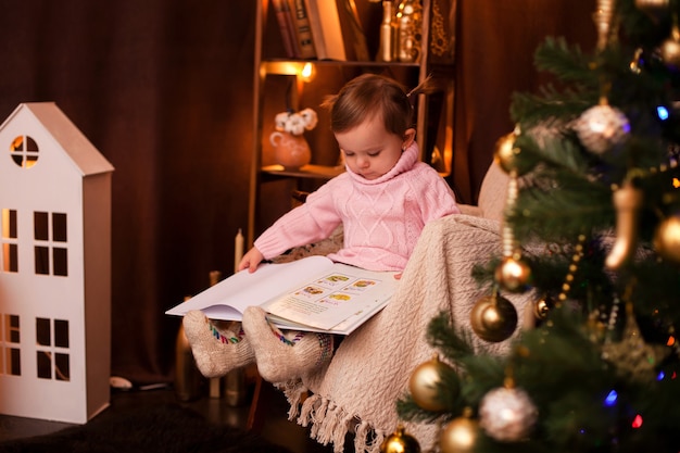 Schattige babymeisje in een trui leest een boek in een kerstinterieur met lichtjes