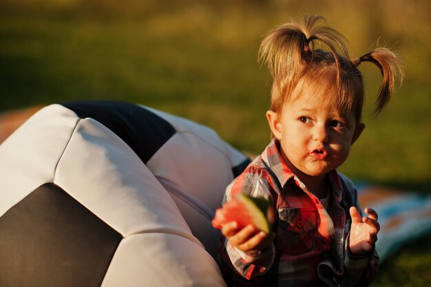 Schattige babymeisje in geruit hemd zit op de poef van de voetbalbal en eet watermeloen