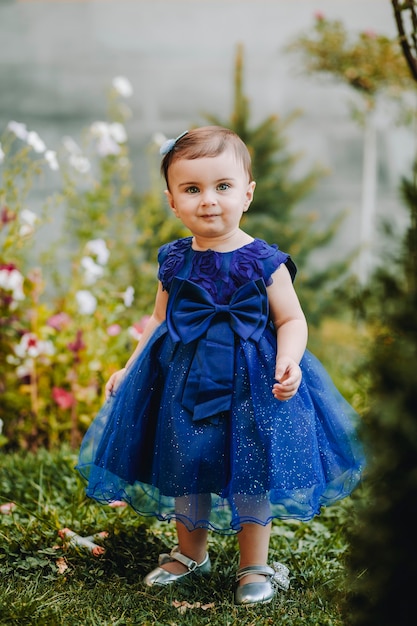 Schattige babymeisje met blauwe jurk staande op het gras in de tuin met veel bloemen