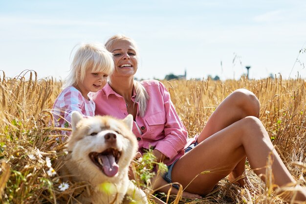 Schattige babymeisje met moeder en hond op tarweveld. Gelukkige jonge familie genieten van tijd samen in de natuur. Moeder, babymeisje en hond husky rusten buiten. saamhorigheid, liefde, geluk concept.