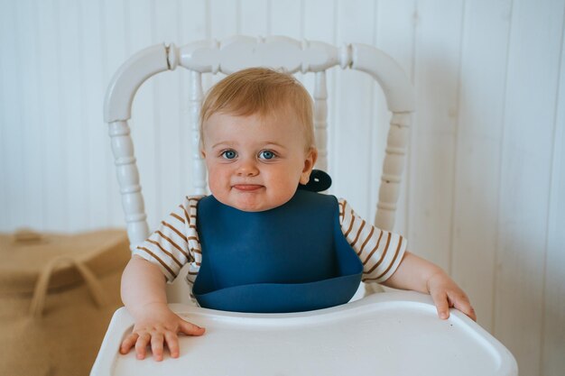 Schattige blanke kleine jongen zit op een voedingsstoel kijkt naar de camera in de keuken en wacht op eten