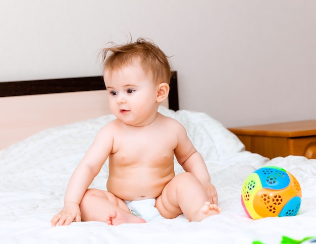 Schattige blonde baby spelen met een bal zittend op het bed in de slaapkamer. happy baby 6 maanden oud spelen met een bal