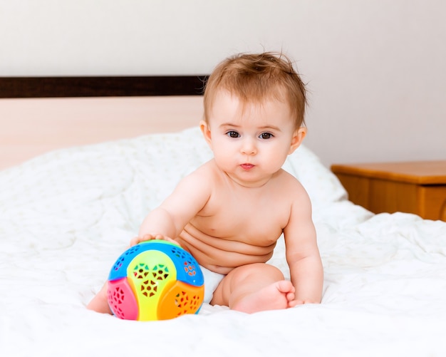 Schattige blonde baby spelen met een bal zittend op het bed in de slaapkamer. happy baby 6 maanden oud spelen met een bal