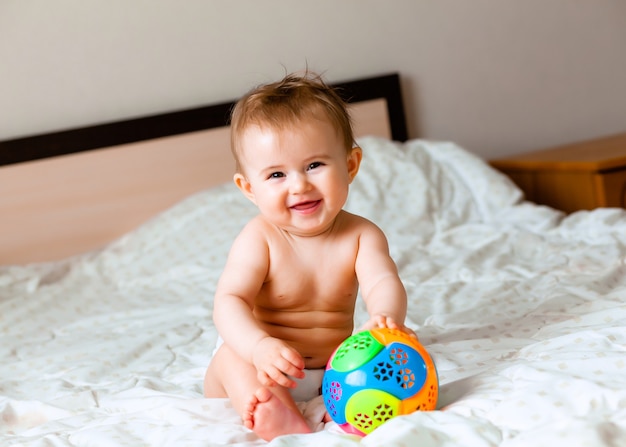 Schattige blonde baby spelen met een bal zittend op het bed in de slaapkamer. happy baby 6 maanden oud spelen met een bal