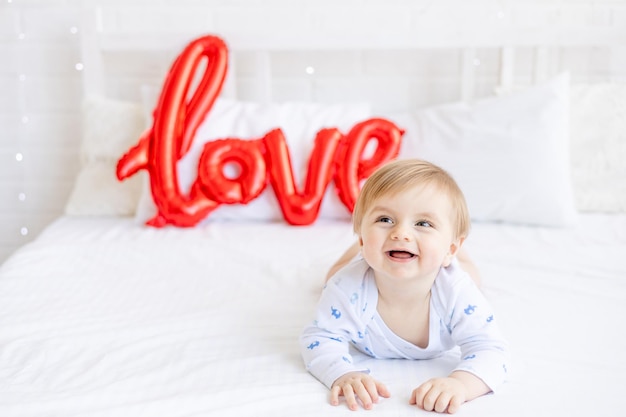 Schattige blonde babyjongen ligt in bed met een grote rode ballon met de inscriptie liefde thuis en lacht in de ochtend het concept van Valentijnsdag of Moederdag