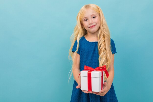 Schattige blonde kind in een jurk met een cadeau in zijn handen op een lichtblauwe achtergrond