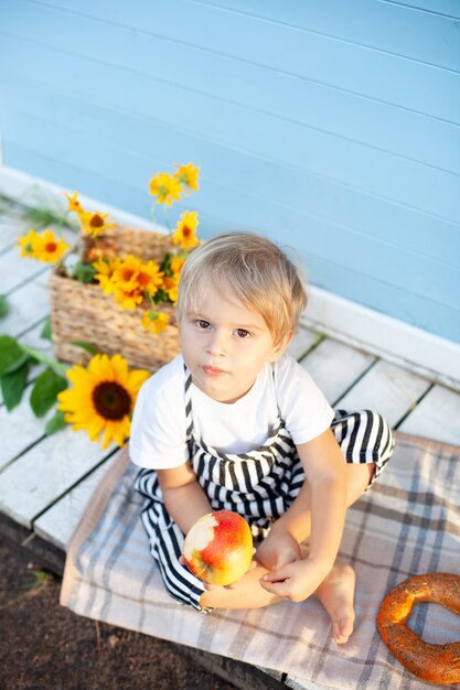 Schattige blonde kleine jongen met een appel en een broodje in zijn handen op de veranda van een houten landhuis op een herfstdag. zomervakantie, openluchtrecreatie. jeugd concept. picnic lunch)