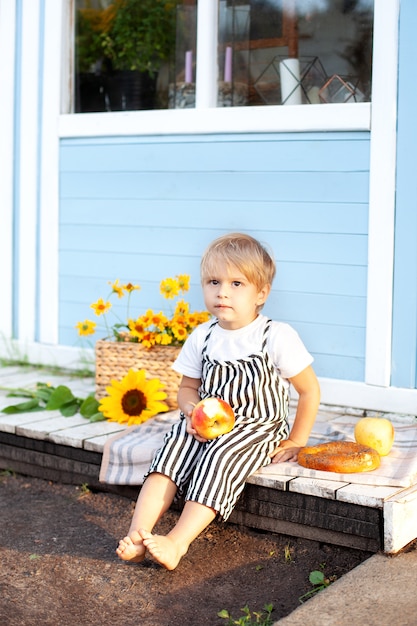 Schattige blonde kleine jongen met een appel en een broodje in zijn handen op de veranda van een houten landhuis op een herfstdag. Zomervakantie, openluchtrecreatie. Jeugd concept. Picnic lunch)