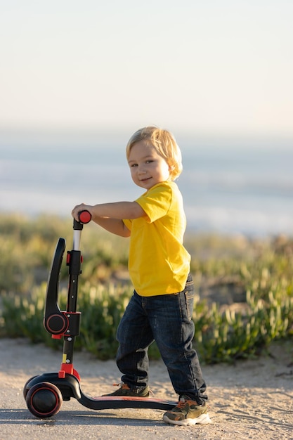 Schattige blonde kleine jongen staat op de weg met een scooter die in de camera kijkt