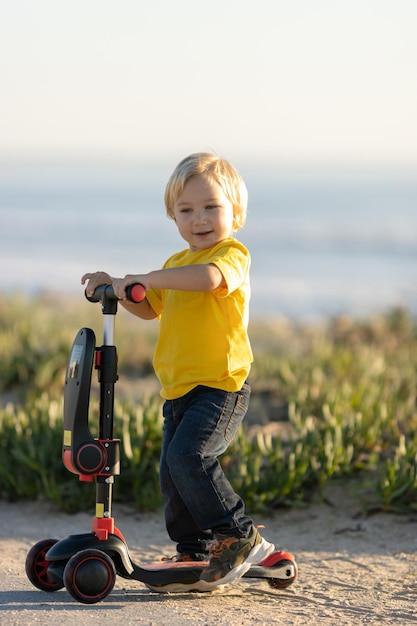 Schattige blonde kleine jongen staat op de weg met een scooter