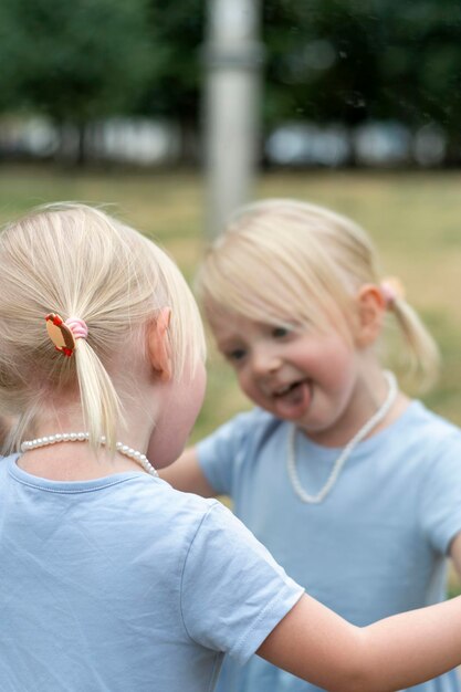 Foto schattige blonde meisje in blauwe jurk toont tong aan haar spiegelbeeld in de spiegel meisje met paardenstaarten