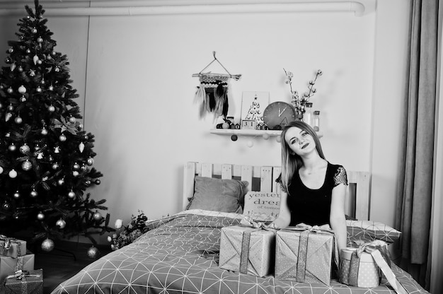Schattige blonde meisje op zwarte jurk liggend op bed met geschenken dozen tegen nieuwjaarsboom in studio. Fijne vakantie.