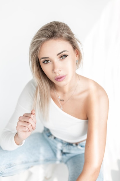 Schattige blonde vrouw in mode witte kleding met tshirt en spijkerbroek zit in studio bij zonlicht