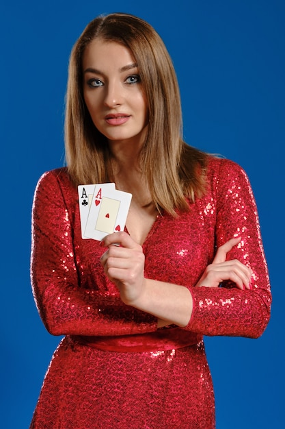 Schattige blonde vrouw met make-up, in rode pailletten jurk toont twee speelkaarten, handen gevouwen, poseren tegen blauwe achtergrond. Gokken entertainment, poker, casino. Detailopname.
