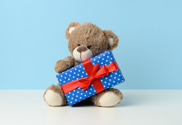 Schattige bruine teddybeer met een doos verpakt in blauw papier en rood zijden lint op witte tafel. Prijs