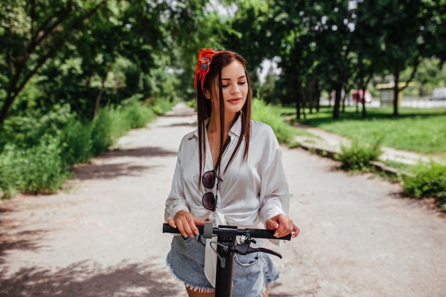 Schattige brunette meisje rijdt op een elektrode scooter in het park dat ze een wit overhemd draagt. concept van eco transport en huur.