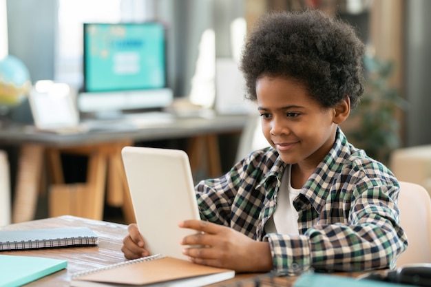 Schattige elementaire schooljongen van Afrikaanse etniciteit zittend aan tafel voor de camera in de woonkamer en kijken naar online educatieve video