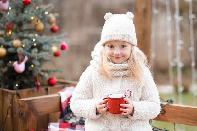 Schattige glimlach meisje van 4-5 jaar oud drinkt warme chocolademelk met rode snoepstok in mok over kerstboom