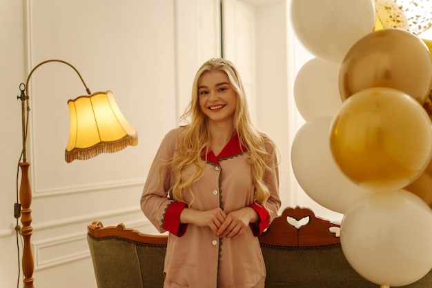 Schattige jonge blanke blonde vrouw in pyjama glimlacht naar de camera met ballonnen op haar verjaardag Vakantieconcept