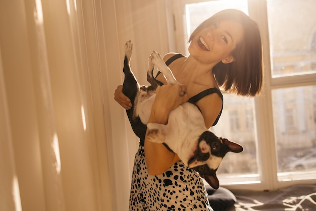 Schattige jonge blanke brunette die zwart-witte Boston Terriër vasthoudt en knuffelt in lichte kamer Liefde voor honden huisdier concept