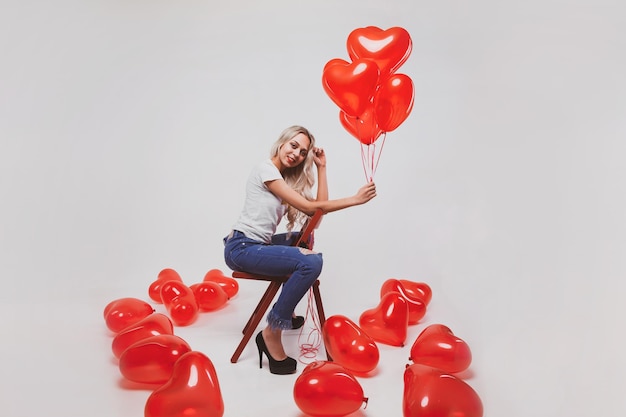 Schattige jonge blonde meisje in jeans en wit t-shirt poseren met hartvormige ballonnen op witte achtergrond Valentijnsdag concept