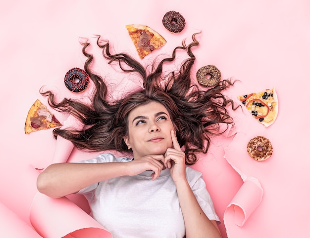 Schattige jonge brunette vrouw op een papier roze achtergrond met pizza en donuts, bovenaanzicht, fastfood concept.