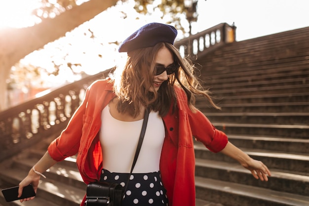 Schattige jonge dame met medium brunette haar blauwe baret stijlvolle zonnebril witte top en rood shirt glimlachend poseren in zonnige herfst stad en naar beneden kijken