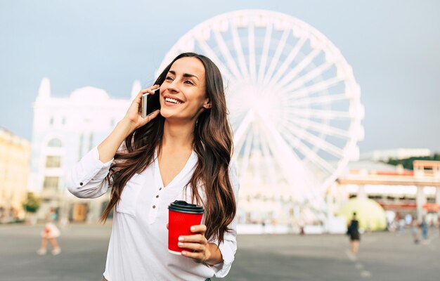 Schattige jonge lachende vrouw praten op smartphone en koffie drinken tijdens het wandelen in de stad