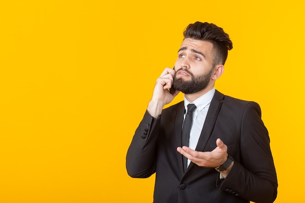 Schattige jonge man met een baard in formele kleding praten aan de telefoon poseren op een gele muur